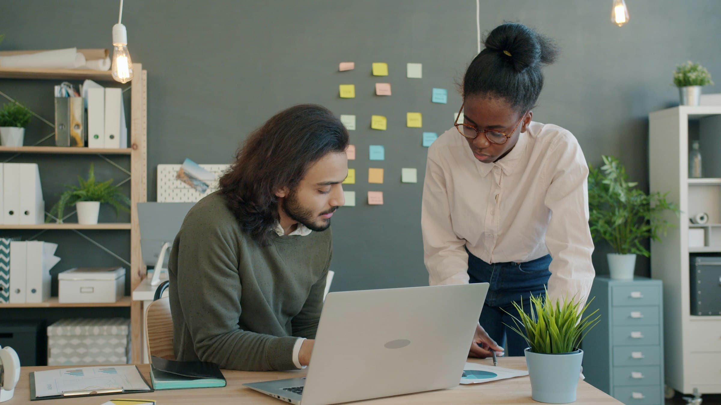 A woman leans over a desk to review work on a laptop with a seated colleague.