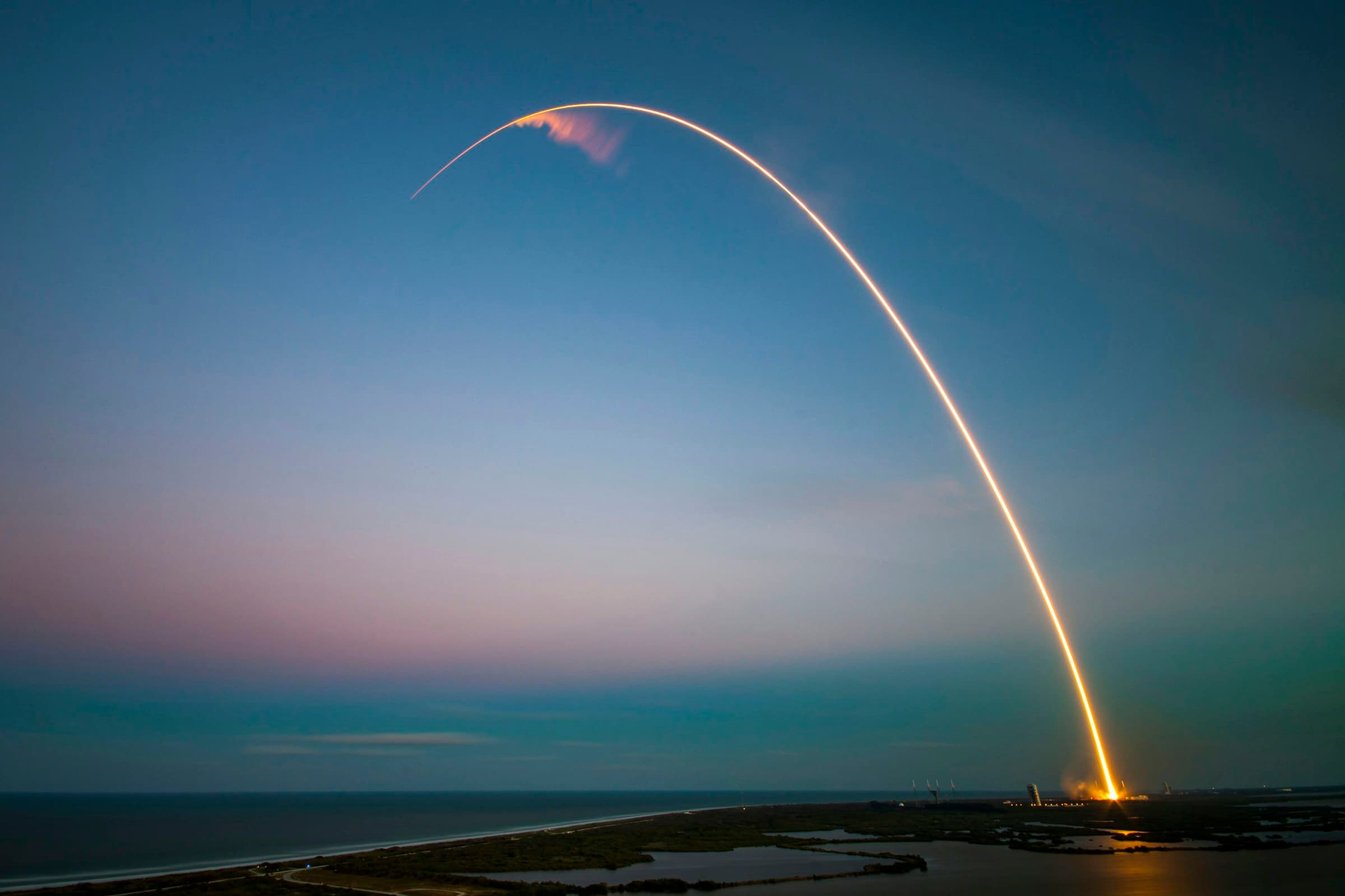 The glowing trail of a rocket launch arcs across a blue dusk sky over the coast.