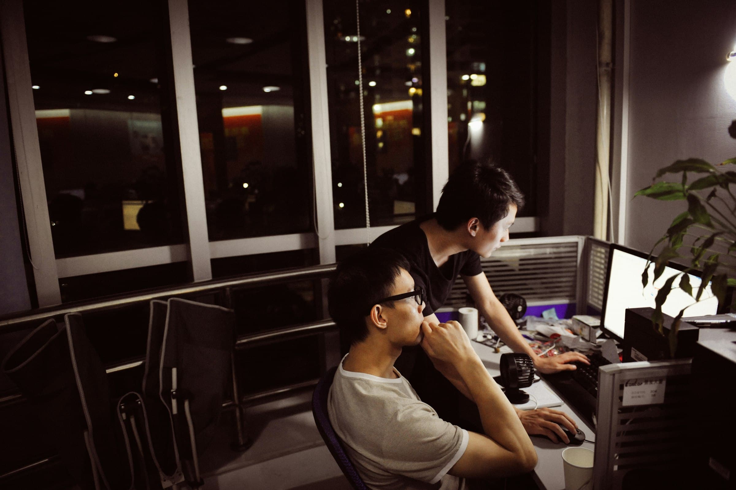 Two engineers work together at a computer in an office at night with city lights outside.