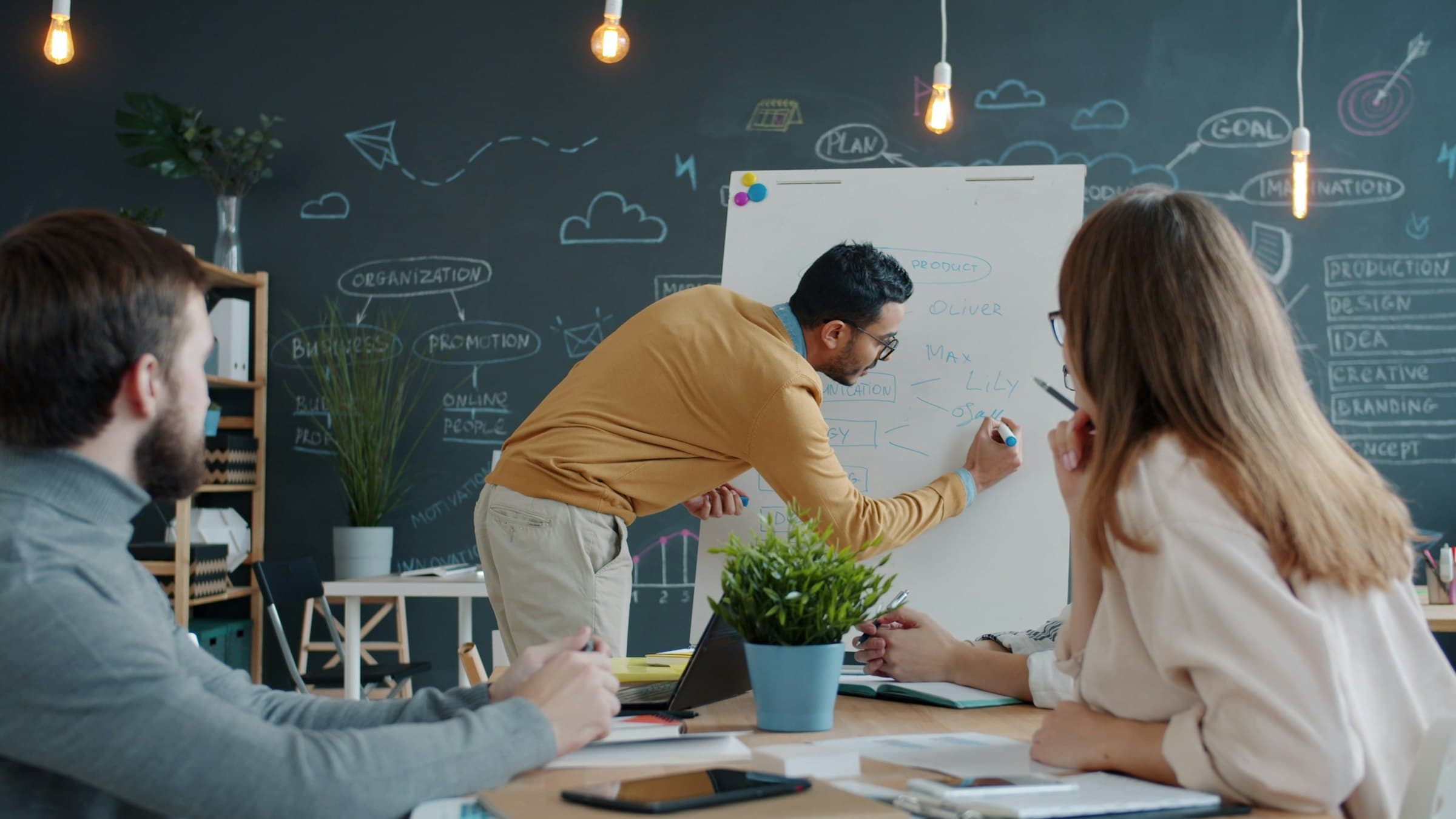 A man sketches a plan on a whiteboard while two colleagues watch from a table.