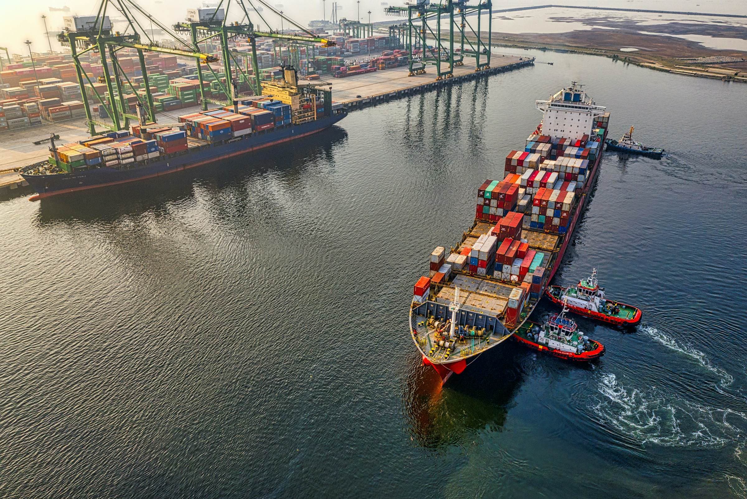An aerial view of a loaded container ship guided out of port by two red tugboats.