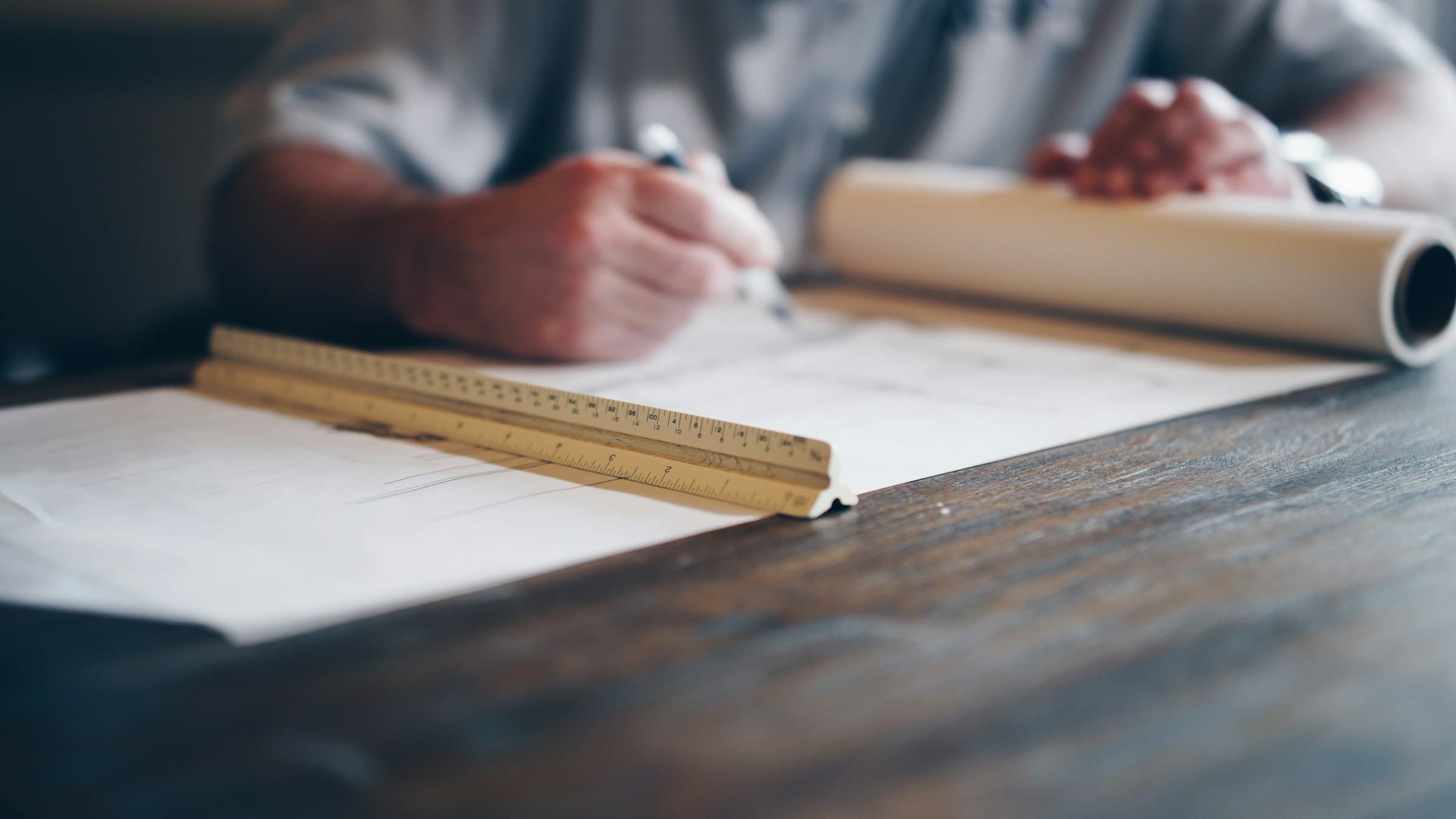 A person drafts on a large sheet with a scale ruler and a rolled blueprint on a wooden table.