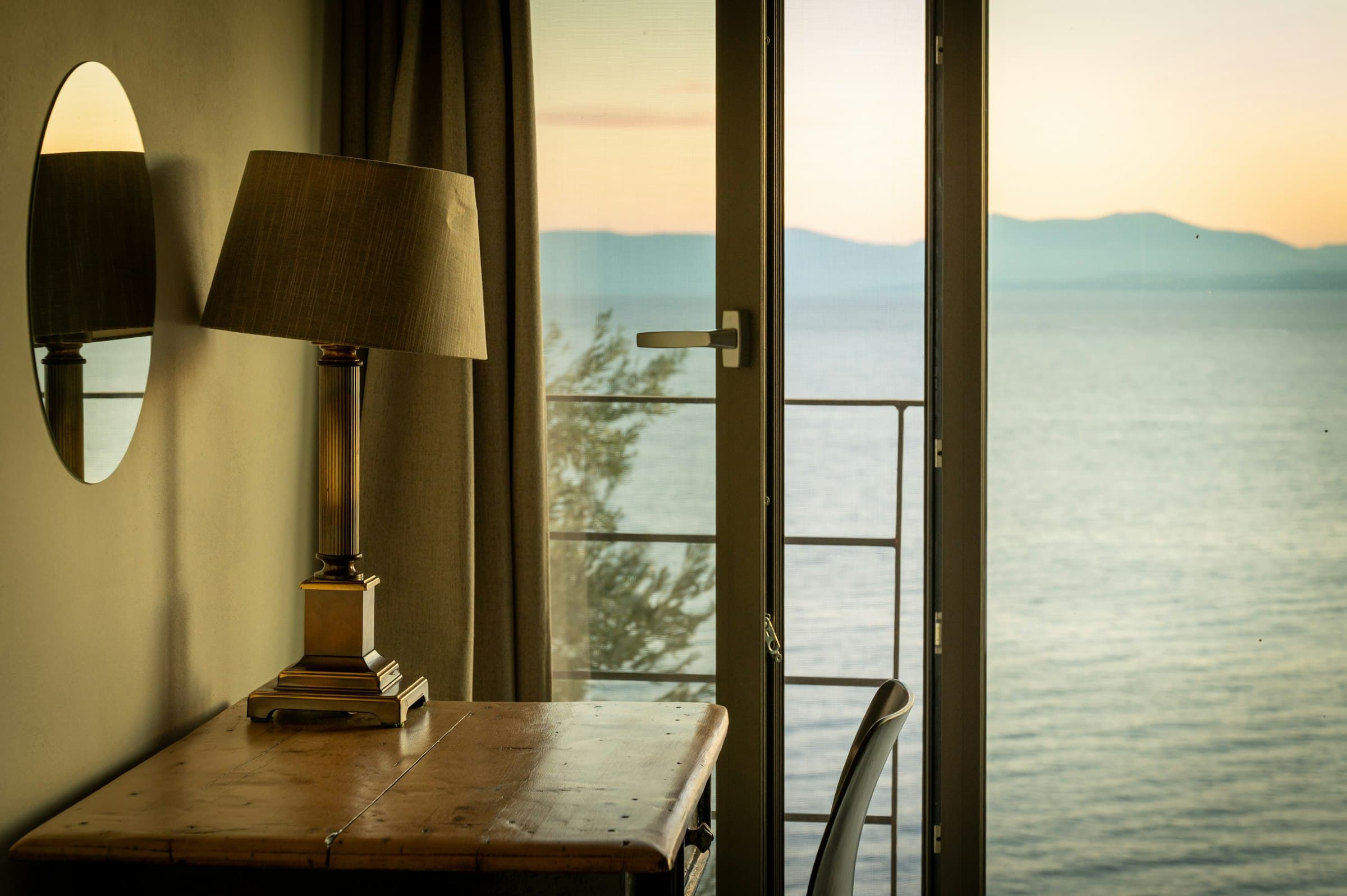 A wooden desk with a brass lamp beside an open balcony door overlooking the sea at dawn.