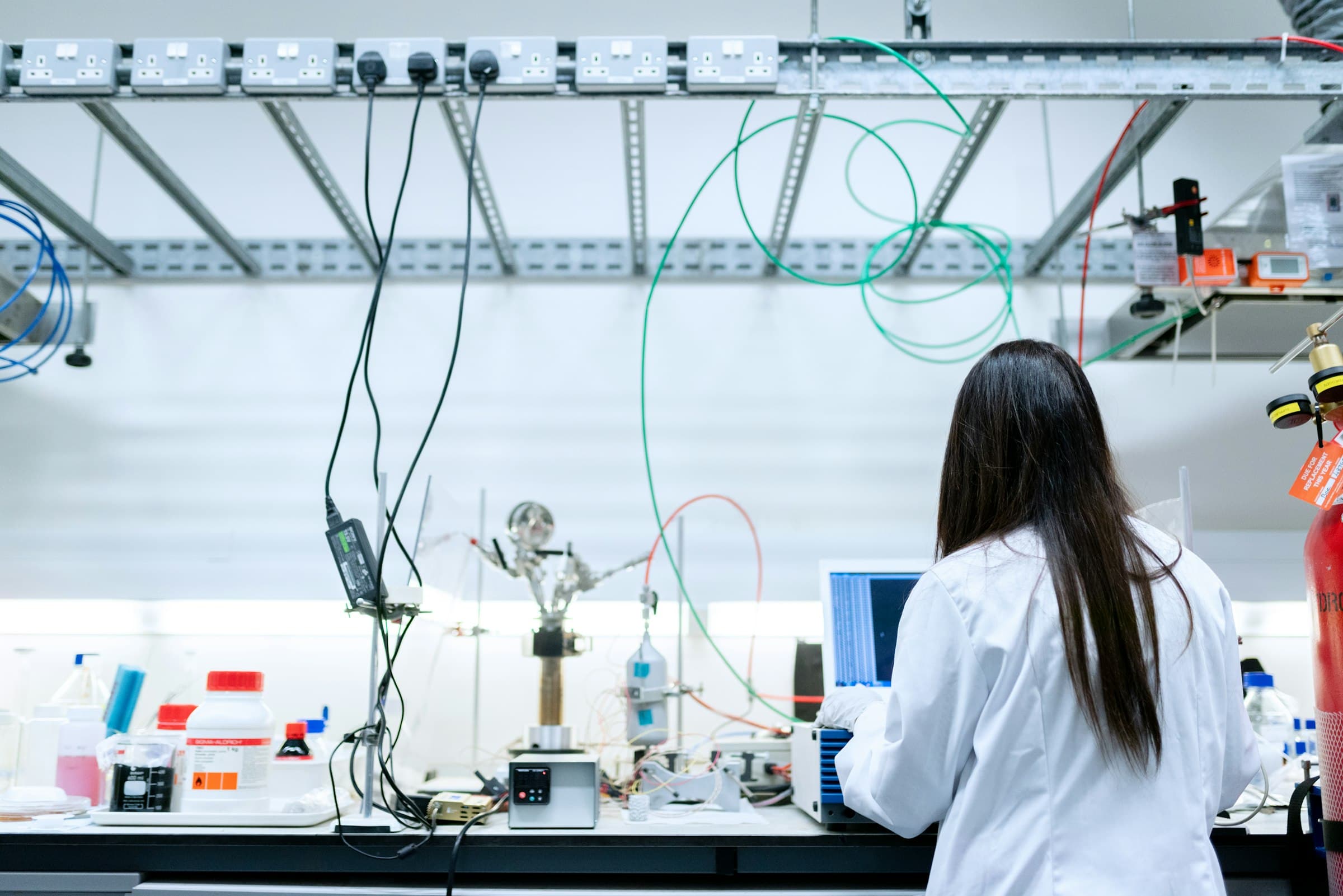 A scientist in a white lab coat works at a lab bench full of instruments and cables.
